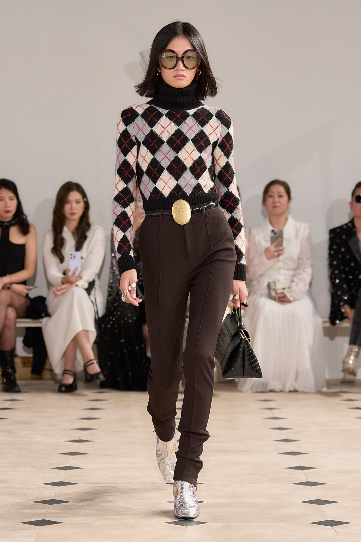 Model walking a fashion runway in a black turtleneck argyle sweater, brown tailored pants, oversized sunglasses, a gold belt buckle, and silver boots.