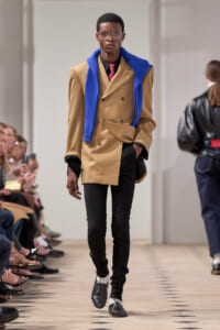 Male model walks a fashion runway in a camel double-breasted coat, blue scarf draped over shoulders, black pants and oxford shoes.