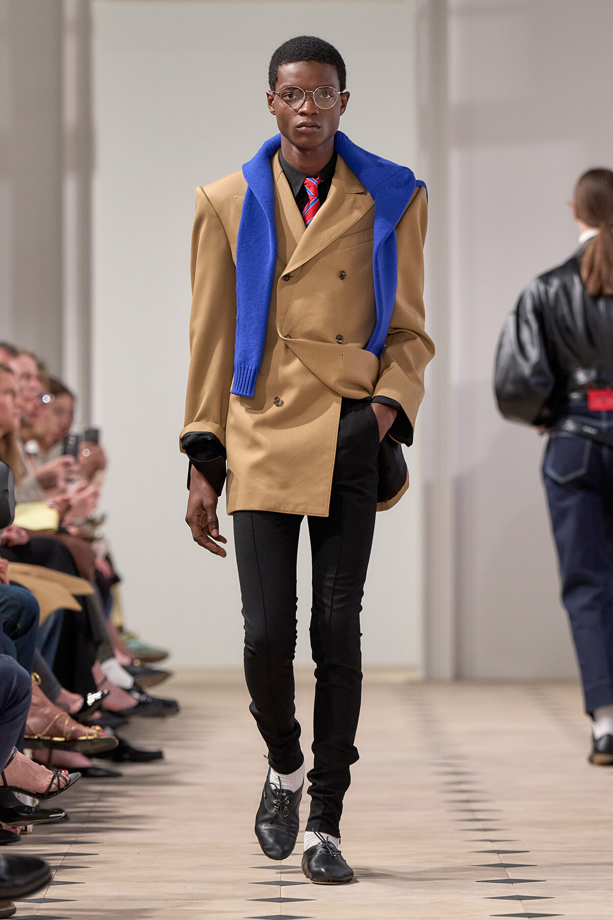 Male model walks a fashion runway in a camel double-breasted coat, blue scarf draped over shoulders, black pants and oxford shoes.