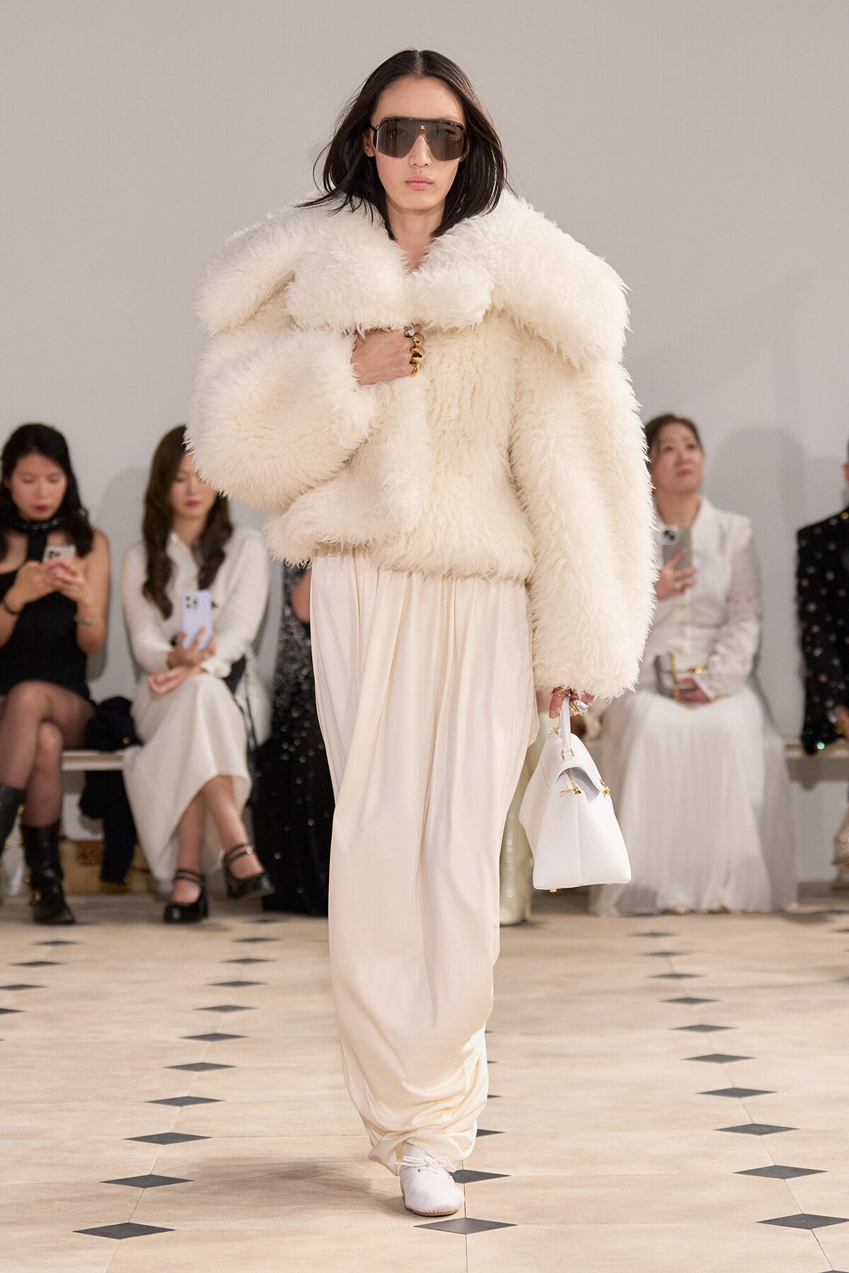 Model on runway wearing an oversized white faux fur coat, cream draped pants, white handbag, and oversized sunglasses.