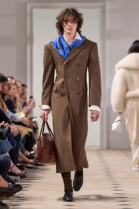 Model on runway wearing a brown double-breasted overcoat, white shirt, blue scarf, and carrying a brown leather bag.