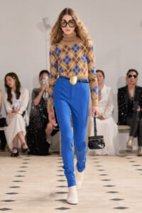 Model walks the runway in a tan-blue argyle sweater, cobalt blue pants, white boots, oversized sunglasses, and a small black handbag.