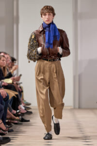 Male model on a fashion runway wearing a brown leather jacket, blue scarf, and tan pleated pants with a left sleeve covered in coins or metallic embellishments.
