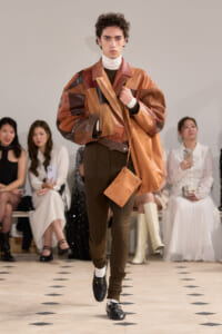 Male model wearing a patchwork brown leather jacket walks the runway, with a tan belt, brown pants, and a matching bag visible beside him.