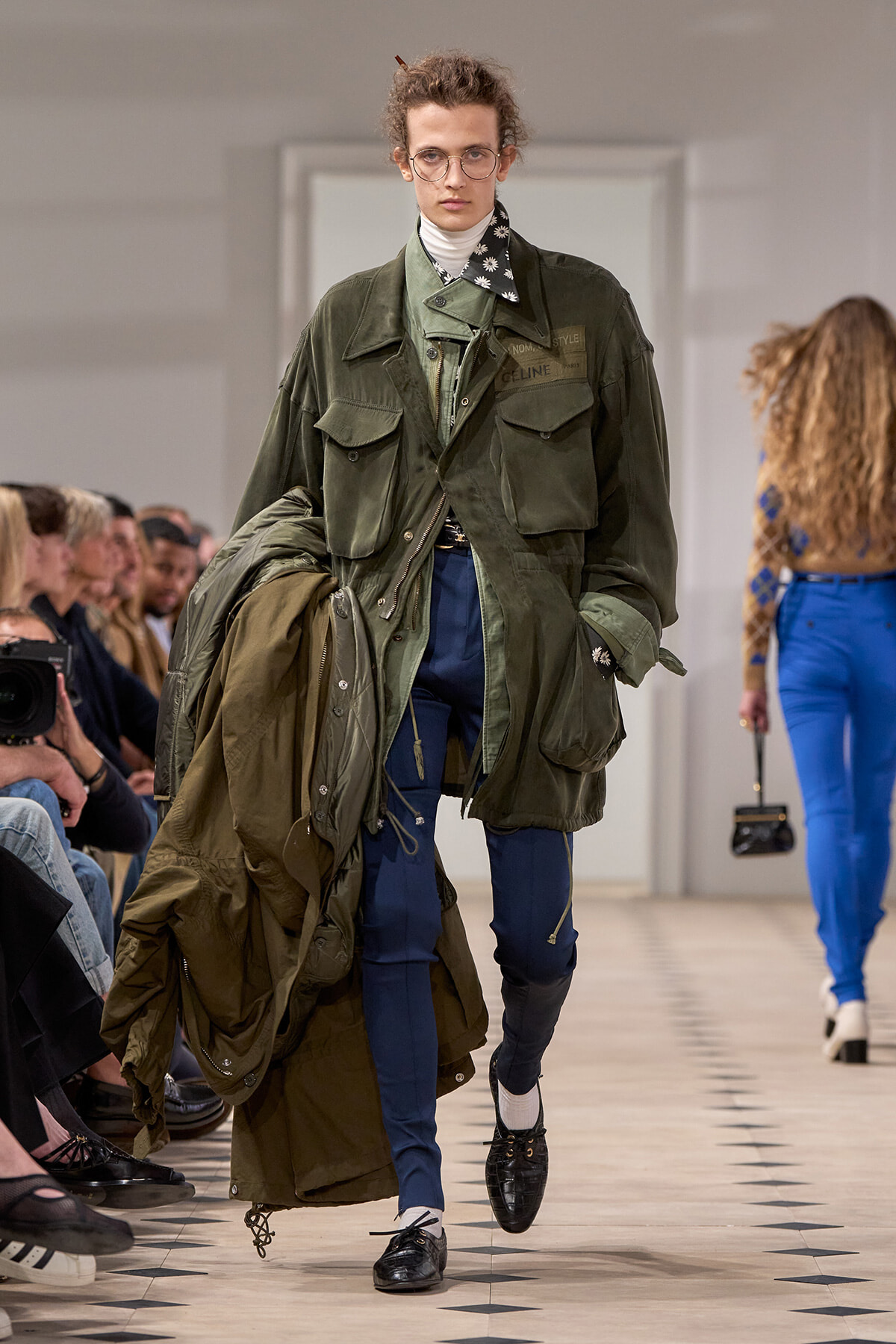 Model walks runway in an oversized olive-green utility jacket with large pockets, paired with blue pants and black shoes, carrying another coat over his arm.