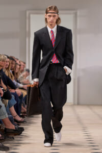 Male runway model in an oversized black suit with a red striped tie, white shirt, and white shoes, carrying a brown patterned tote bag as audience watches.