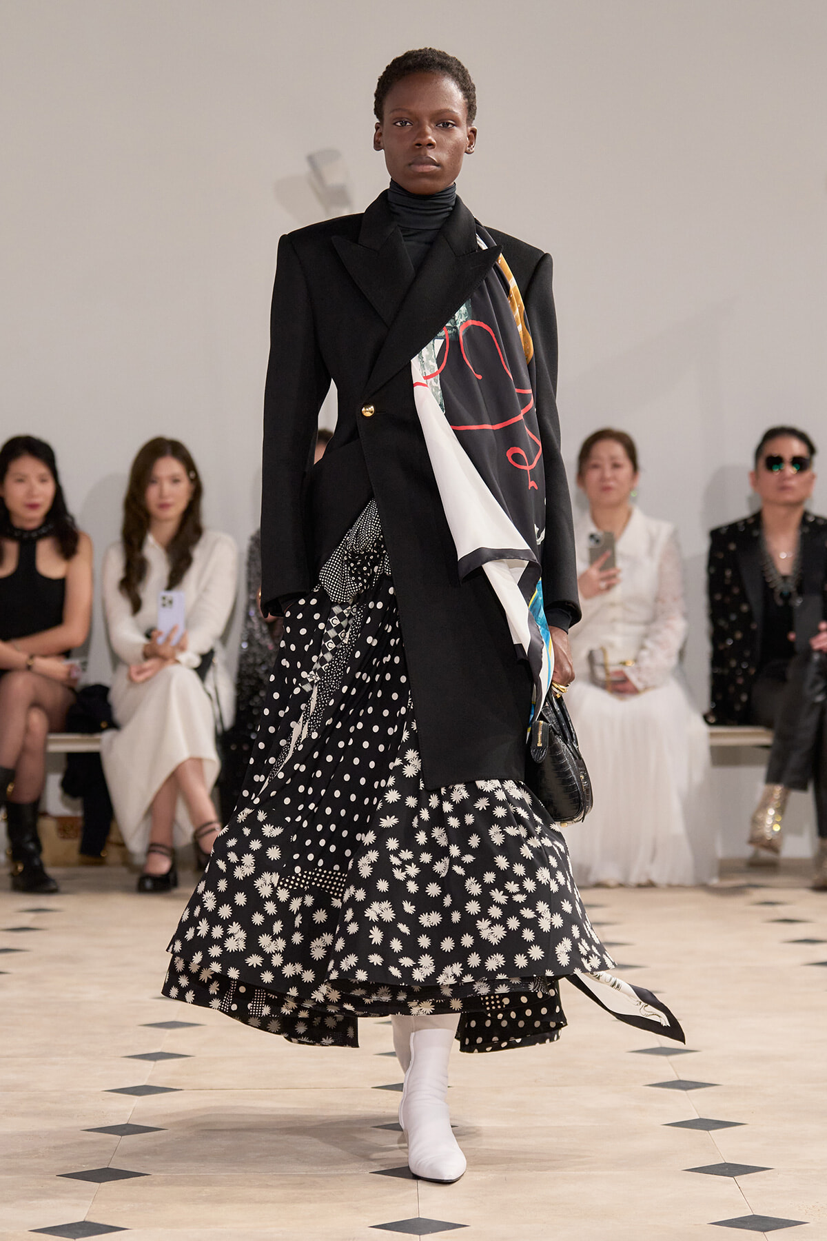 Runway model walking center stage in a black blazer over a patterned black-and-white skirt and white boots, with a colorful scarf draped over the shoulder.