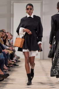 Model walks a runway in a black knit dress with white collar and cuffs, tan handbag, and round sunglasses.