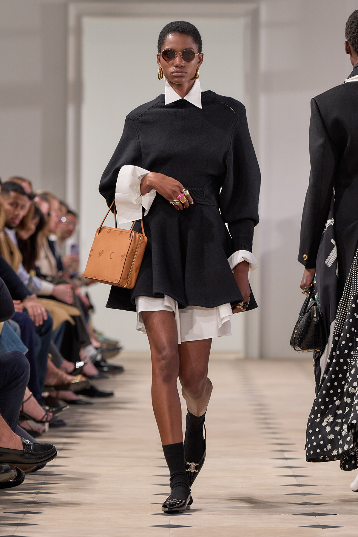 Model walks a runway in a black knit dress with white collar and cuffs, tan handbag, and round sunglasses.