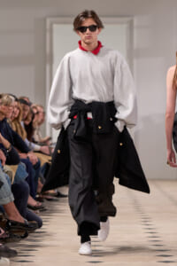 Male model walking a fashion runway in an oversized light gray sweatshirt with a red collar, black draped wrap tied at the waist, and wide black pants; wearing white sneakers and dark sunglasses.