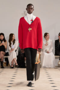 Male model on a runway wearing a bright red V-neck sweater over a white turtleneck, black trousers, and black shoes, carrying a large brown tote bag with gold handles; layered necklaces visible.