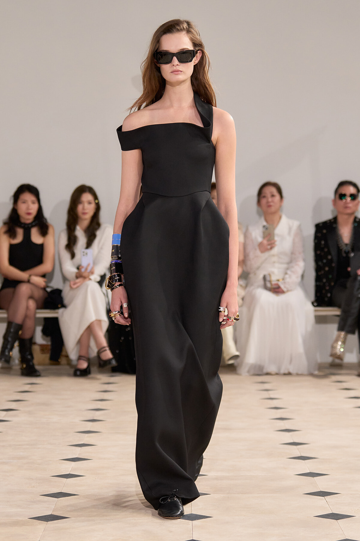 Model walks a black off-shoulder gown on a runway, wearing large dark sunglasses, with seated spectators in background.