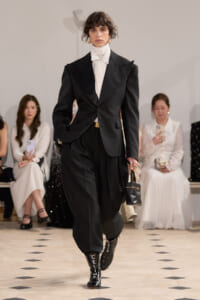 Model on a runway wearing an oversized black power suit with a white turtleneck, carrying a small black handbag; audience seated in the background.
