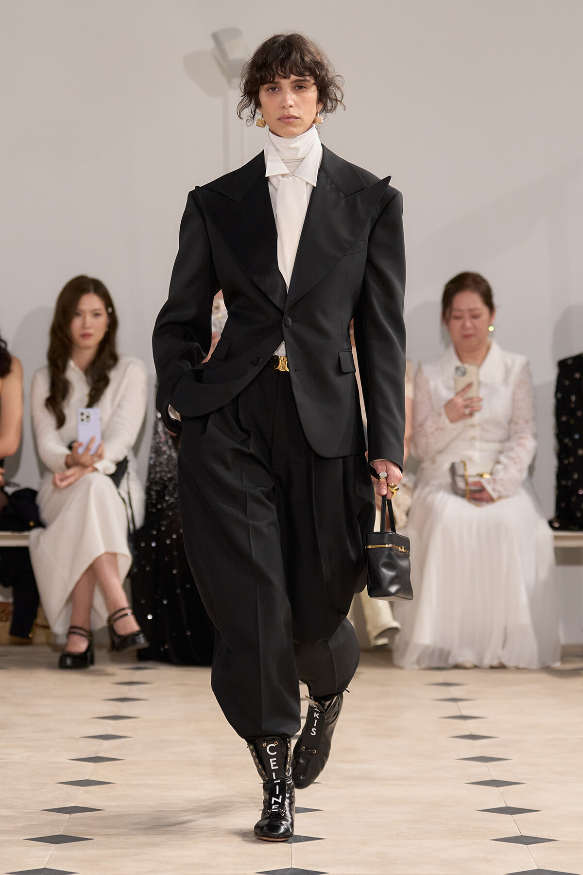 Model on a runway wearing an oversized black power suit with a white turtleneck, carrying a small black handbag; audience seated in the background.