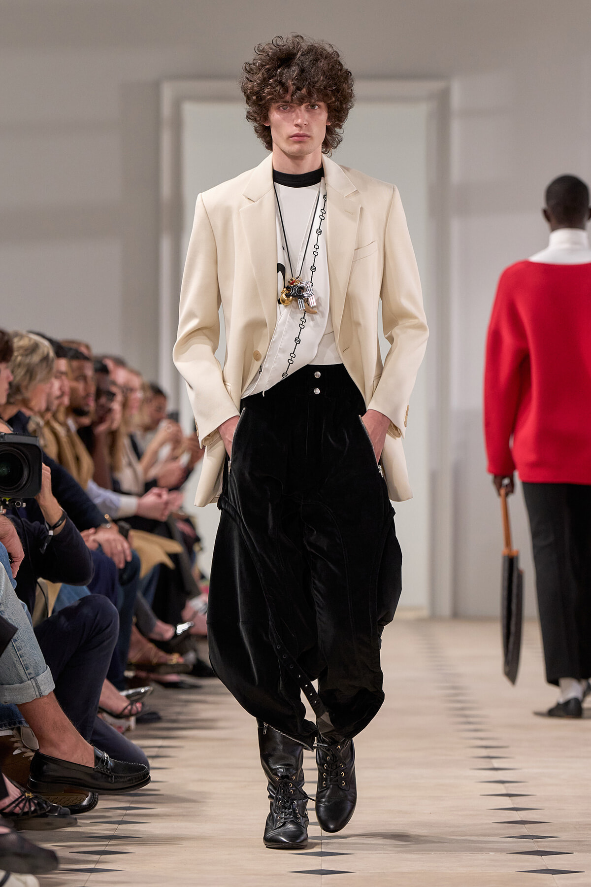 Male model on a fashion runway wearing a beige blazer, white shirt, black loose trousers, and black lace-up boots with layered necklaces.