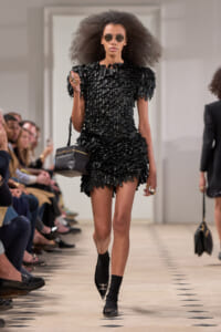Model walking a fashion show wearing a black textured fringe mini dress; large afro, sunglasses, and a small black handbag with gold accents as she strides down the runway.