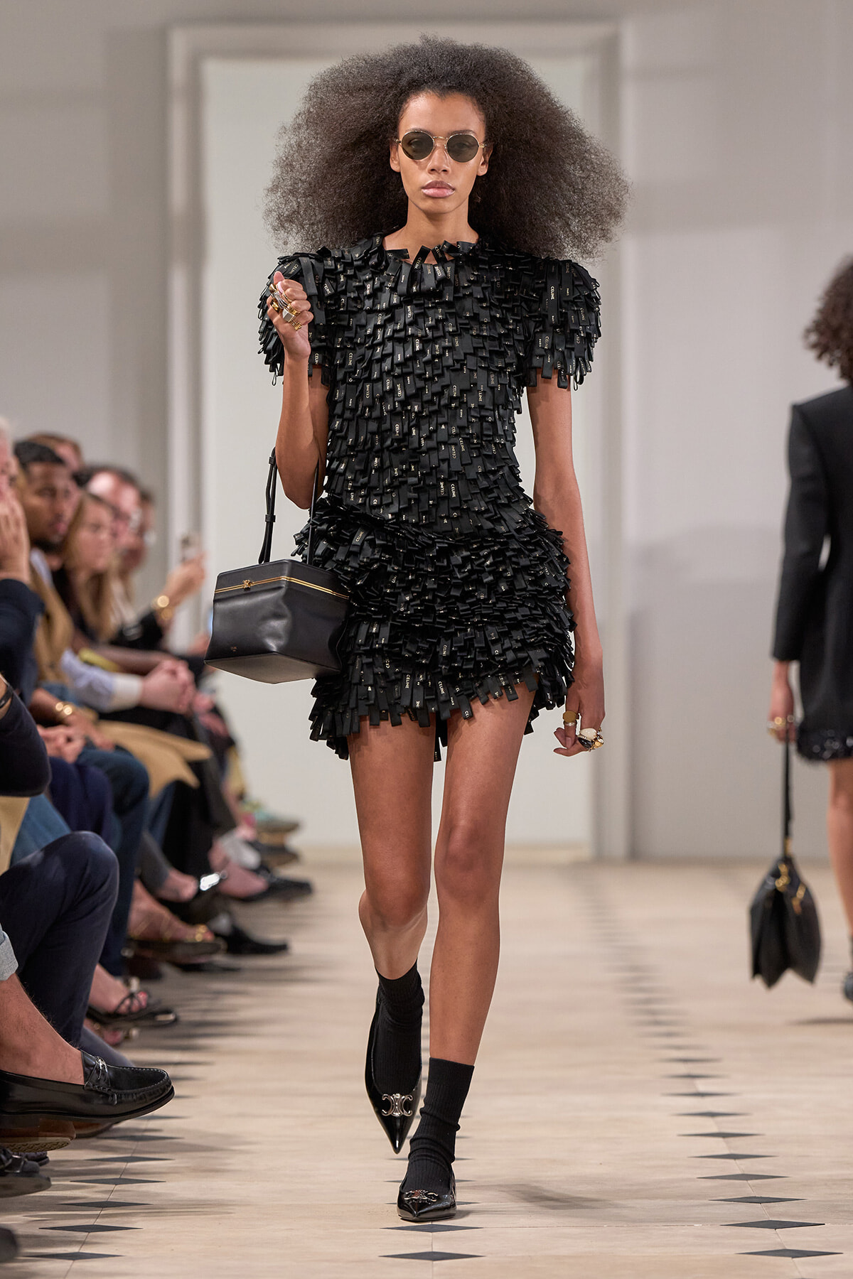 Model walking a fashion show wearing a black textured fringe mini dress; large afro, sunglasses, and a small black handbag with gold accents as she strides down the runway.
