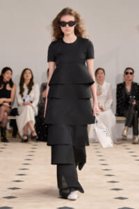 Model walking a runway in a black tiered dress with short sleeves and oversized sunglasses, carrying a white tote bag.