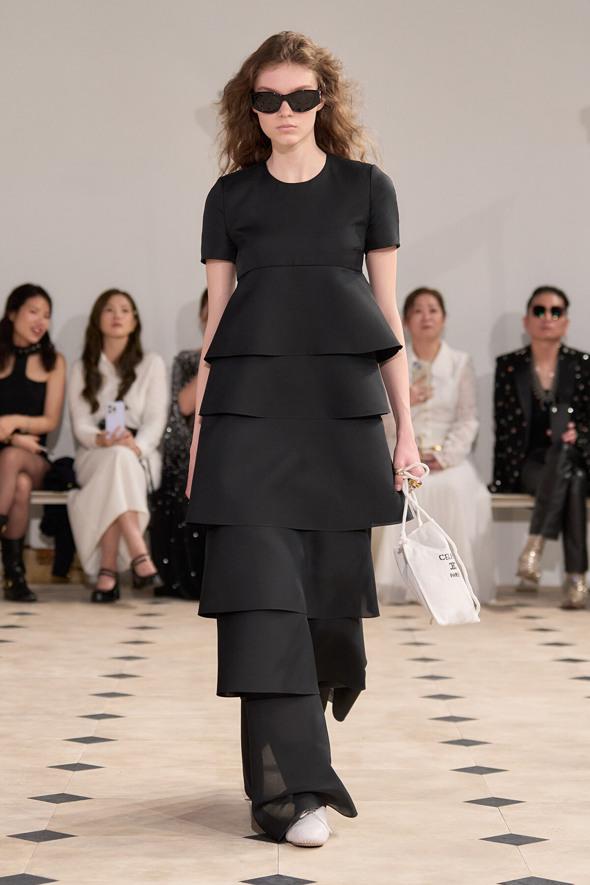 Model walking a runway in a black tiered dress with short sleeves and oversized sunglasses, carrying a white tote bag.