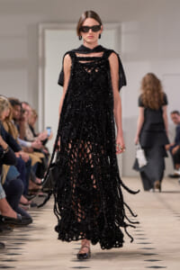 Model on a runway wearing a black sequined dress with a netted, fringed overlay and capelet, sunglasses, and black flats.