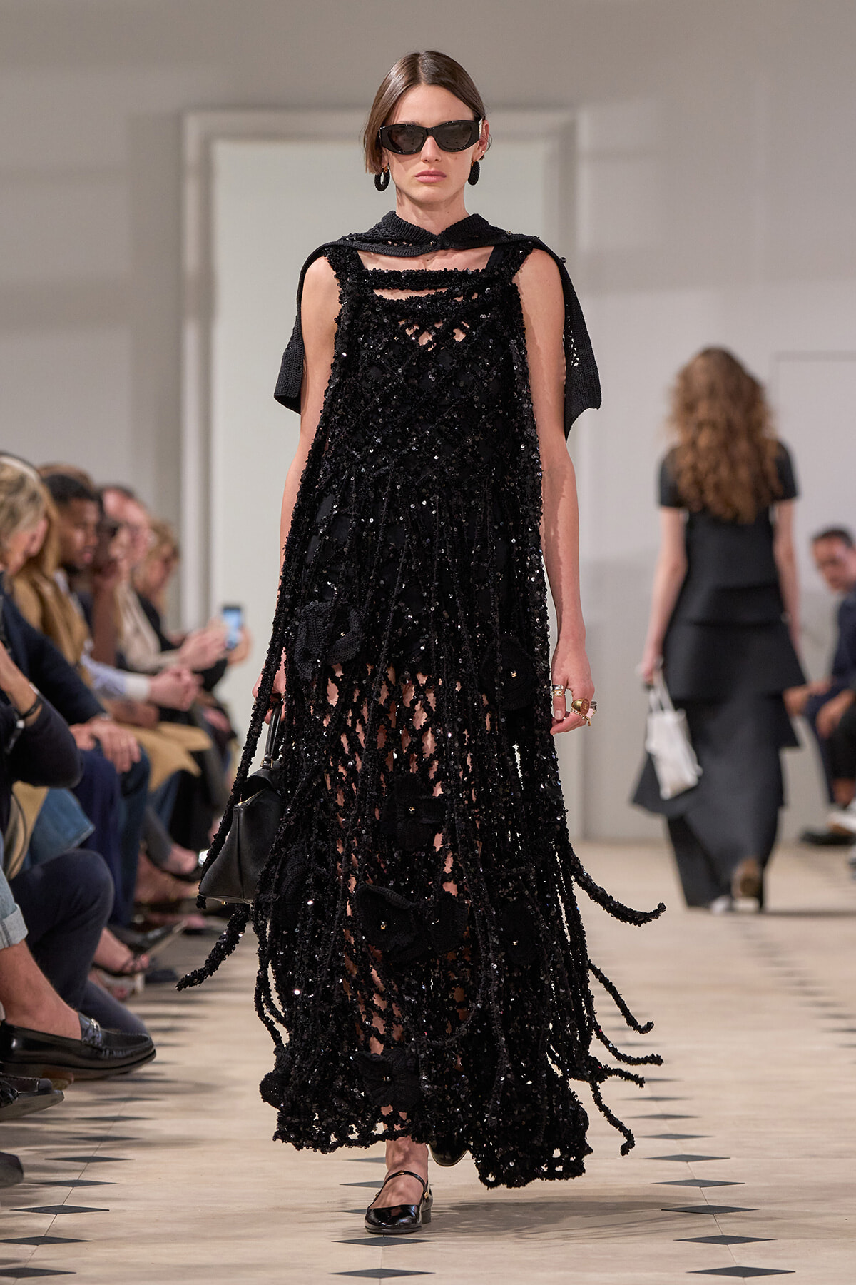 Model on a runway wearing a black sequined dress with a netted, fringed overlay and capelet, sunglasses, and black flats.