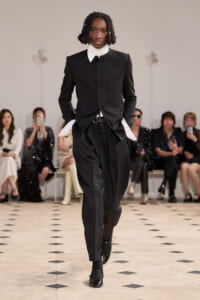Male model walks a runway in a black tailored jacket with white cuffed sleeves and matching trousers, audience seated in the background.