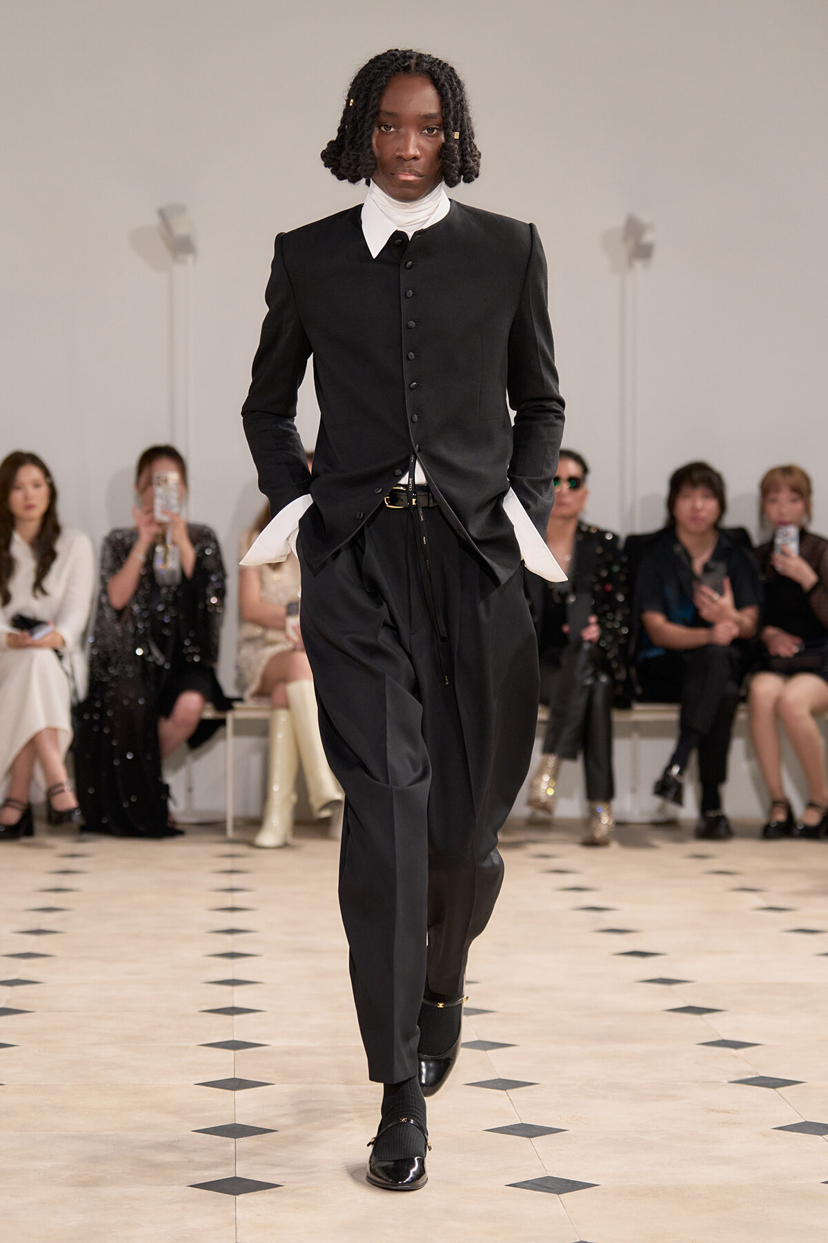 Male model walks a runway in a black tailored jacket with white cuffed sleeves and matching trousers, audience seated in the background.