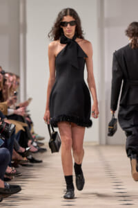 Model walks the runway in a black halter-neck mini dress with a large bow at the neck and a fringed hem, paired with black sunglasses and a small black handbag.