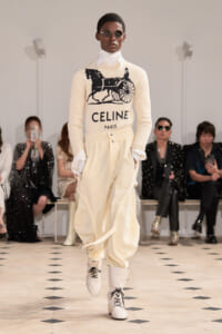 Male model on a runway wearing a cream CELINE sweater with a horse-drawn carriage graphic and 'CELINE PARIS' logo, paired with off-white cargo pants and white lace-up boots.