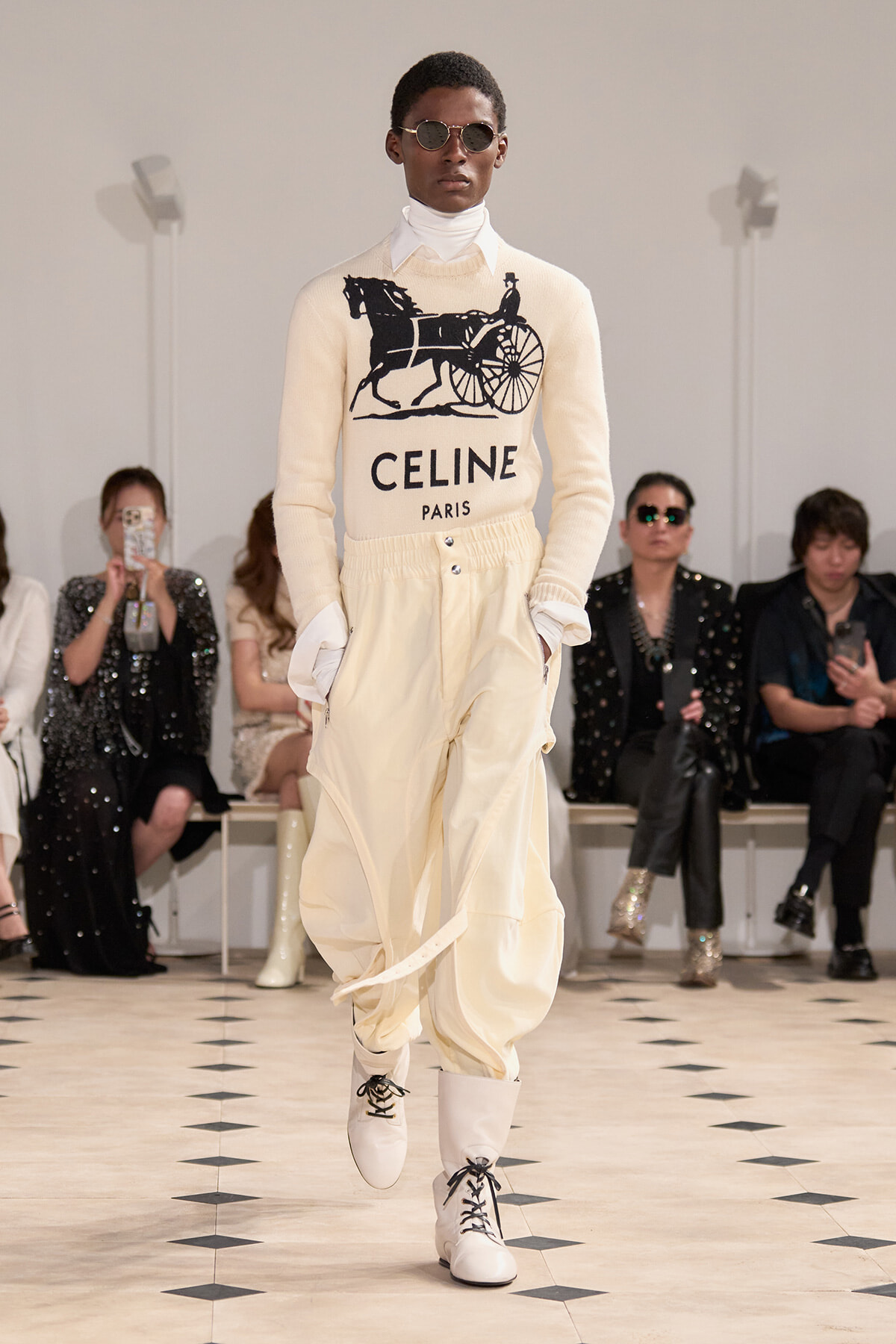 Male model on a runway wearing a cream CELINE sweater with a horse-drawn carriage graphic and 'CELINE PARIS' logo, paired with off-white cargo pants and white lace-up boots.
