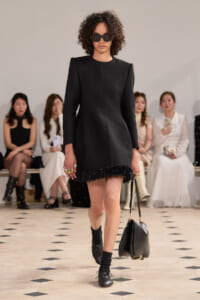 Model walks a runway in a black structured mini dress with a feathered fringe hem, black sunglasses, and a black shoulder bag.