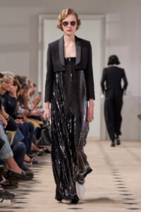 Model on a runway wearing a black sequined strapless gown with a cropped black blazer, round sunglasses, and a small black handbag.