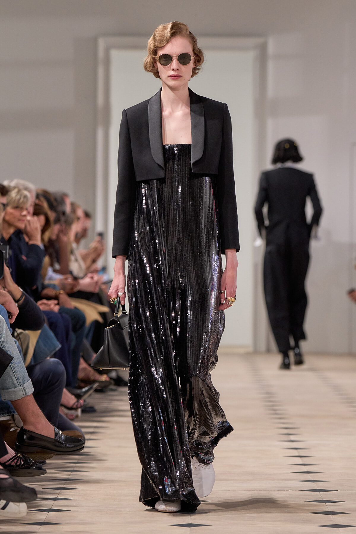 Model on a runway wearing a black sequined strapless gown with a cropped black blazer, round sunglasses, and a small black handbag.