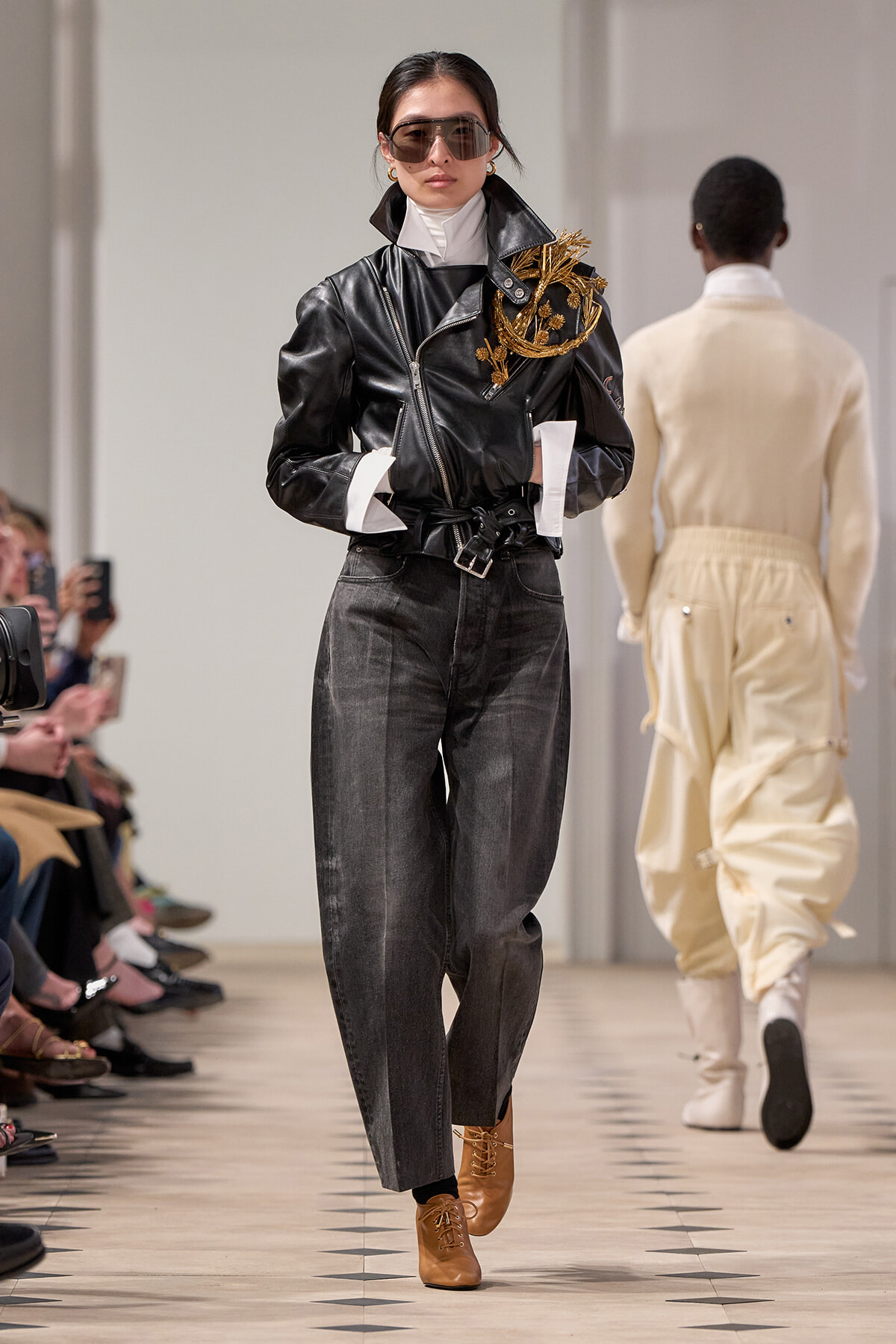 Model walking the runway in a black leather jacket with gold embroidery, white turtleneck, dark jeans, and tan boots with oversized sunglasses.