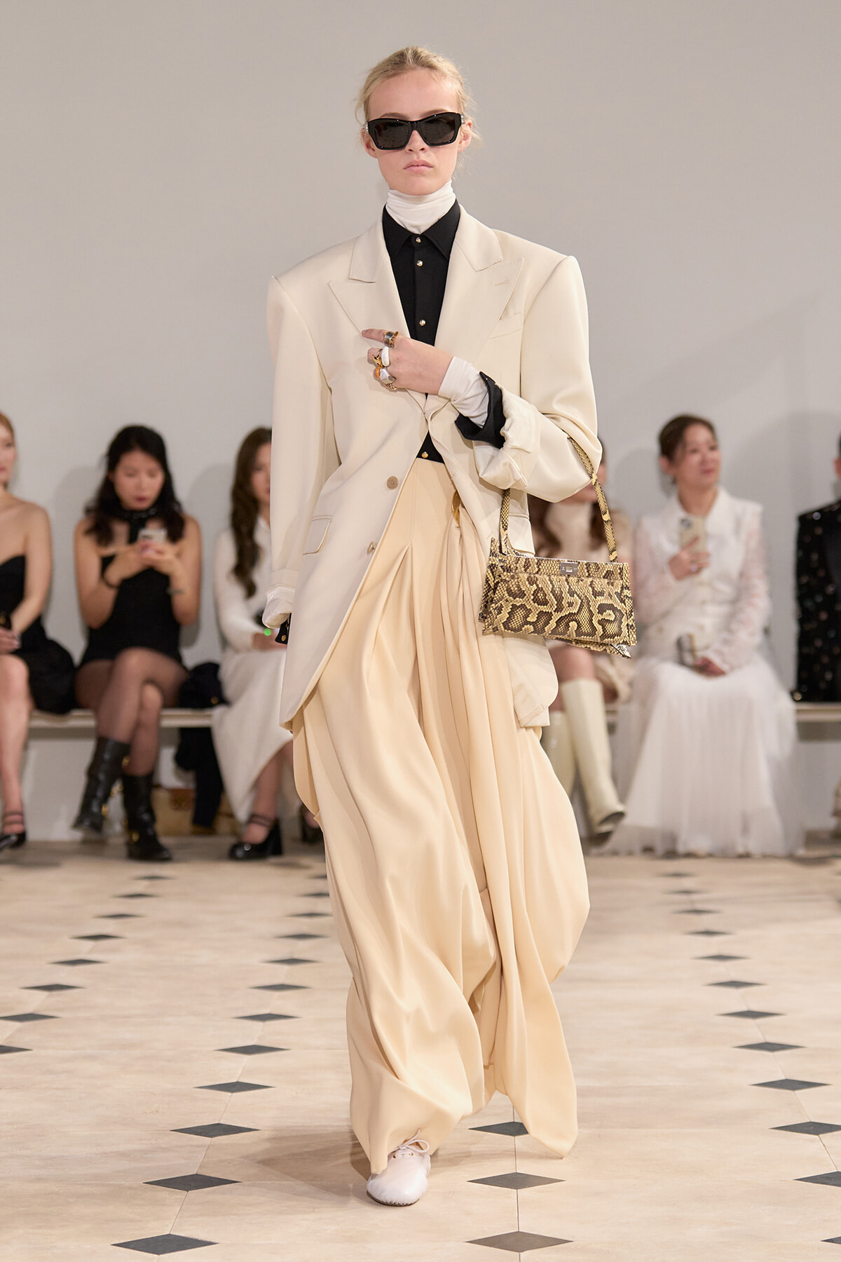 Model walking a fashion runway in a cream oversized blazer over a black turtleneck and wide cream trousers, accessorized with black sunglasses and a snakeskin handbag.