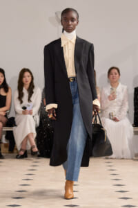 Model walking the runway in a long black blazer over a cream turtleneck and shirt, blue jeans, tan boots, and carrying a black handbag.