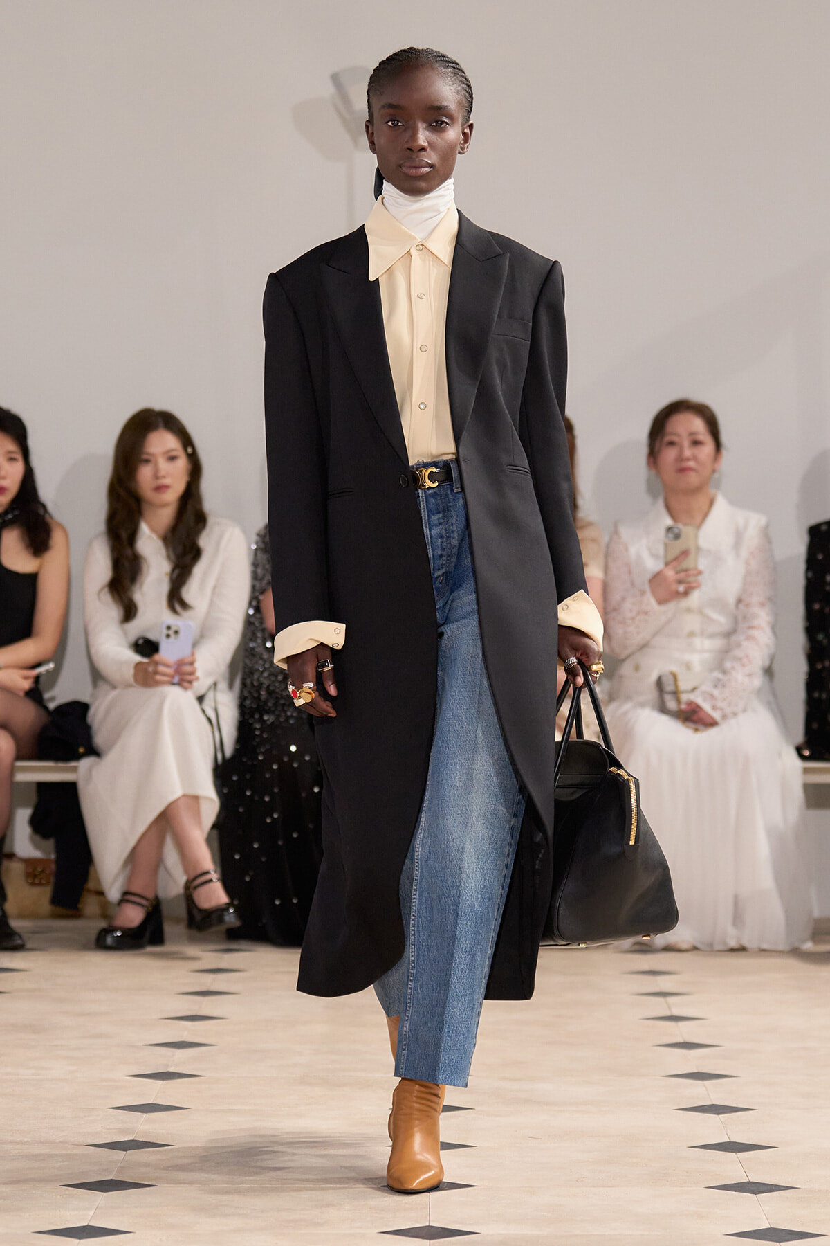 Model walking the runway in a long black blazer over a cream turtleneck and shirt, blue jeans, tan boots, and carrying a black handbag.