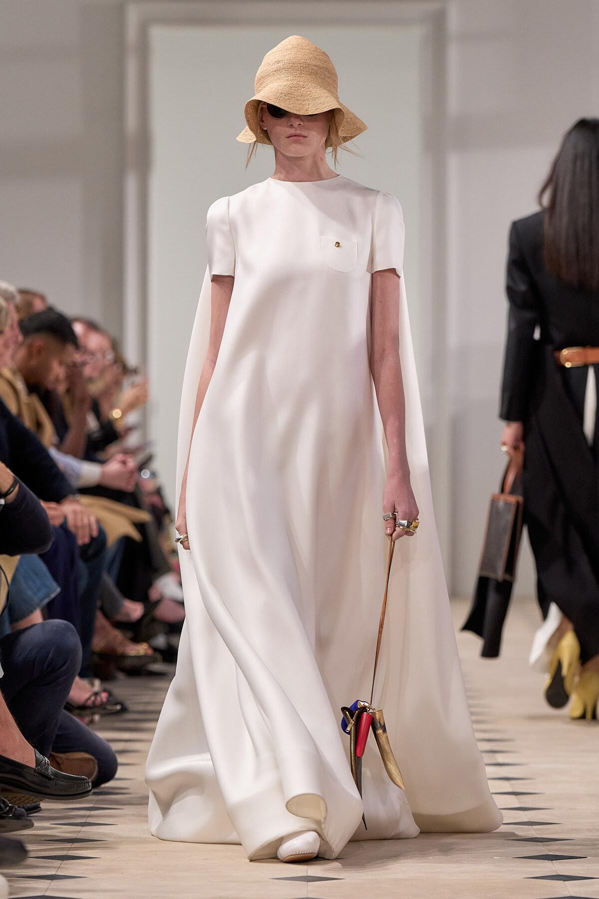 Model on a fashion runway in a white floor-length cape dress with a wide straw hat and sunglasses, carrying a small handbag by a long strap.