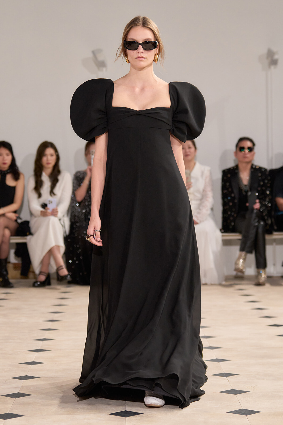 Model on a runway in a black floor-length gown with oversized puff sleeves and dark sunglasses, audience in the background.