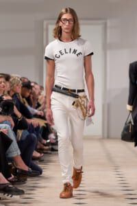 Male model on a runway in a white Celine t-shirt, white jeans, brown boots, and a black belt with gold chains.