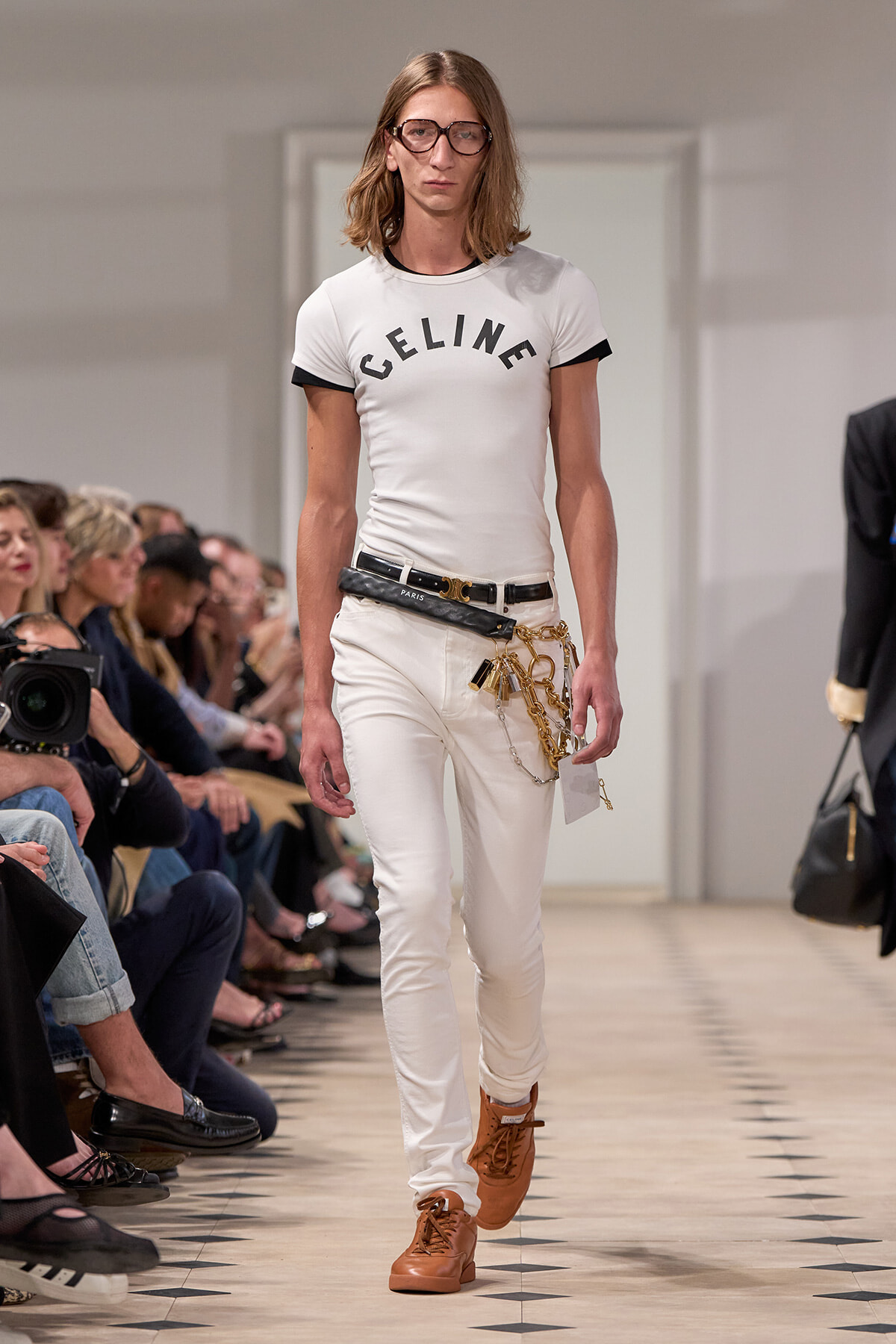 Male model on a runway in a white Celine t-shirt, white jeans, brown boots, and a black belt with gold chains.