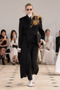 Runway model in a black double-breasted suit with a gold twig shoulder ornament, white turtleneck, and white boots.