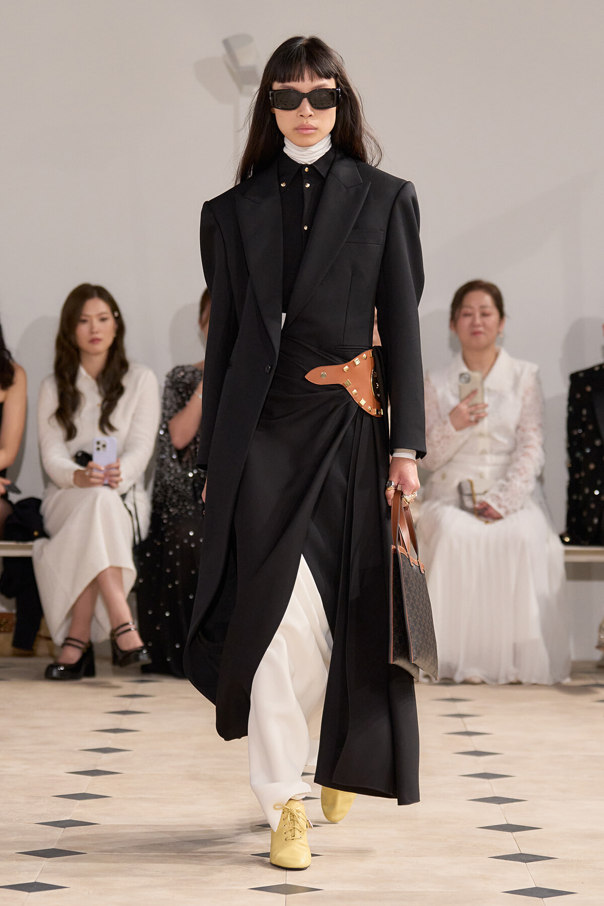 Model walks a fashion show in an oversized black blazer over a draped black dress with white pants, a tan studded belt, and yellow boots, wearing dark sunglasses.