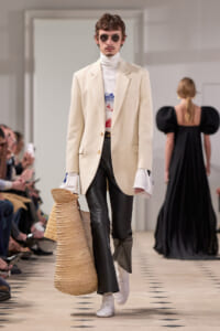 Male fashion model on a runway in a cream blazer, white turtleneck, graphic tee, black leather pants, white sneakers, round sunglasses, carrying a large woven tote.