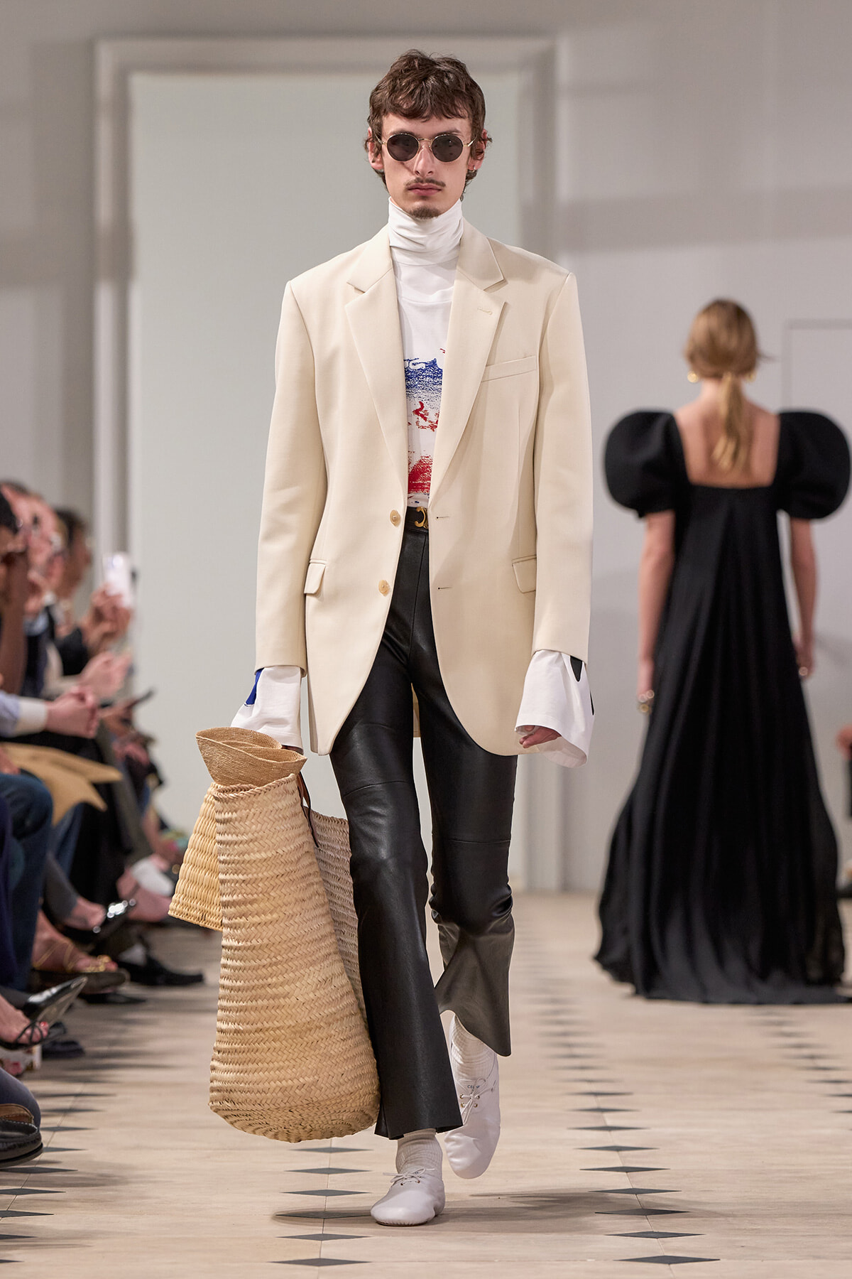 Male fashion model on a runway in a cream blazer, white turtleneck, graphic tee, black leather pants, white sneakers, round sunglasses, carrying a large woven tote.