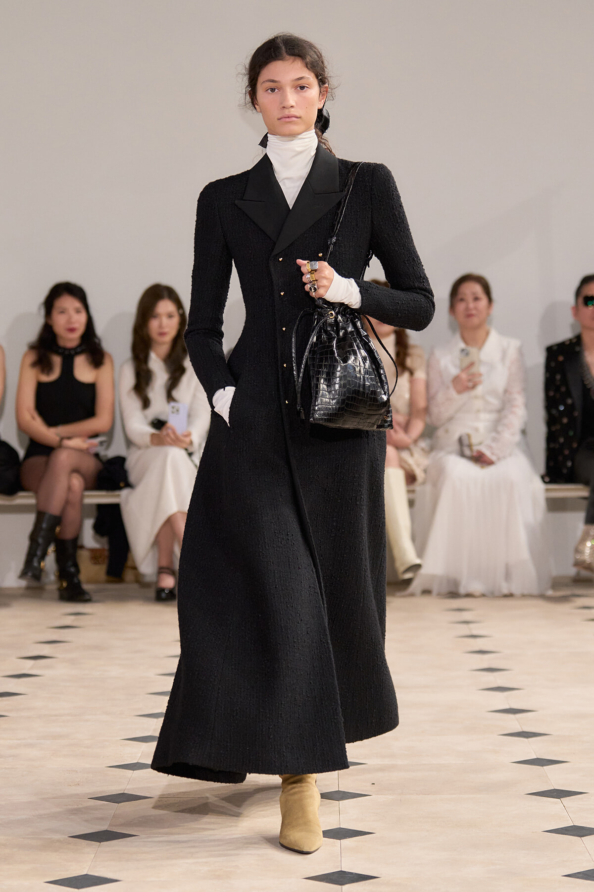Model walks a runway in a long black textured coat with wide lapels, white turtleneck, and beige ankle boots, carrying a black croc-embossed bag.