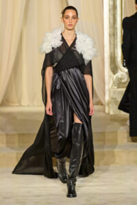 Model on a runway wearing a black satin wrap dress with dramatic white feathered shoulder accents and knee-high black boots.