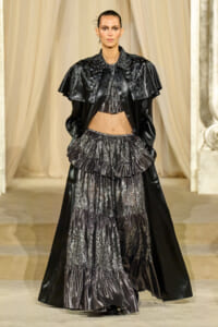 Model walks the runway in a black metallic, tiered gown with lace panels and a dramatic cape-like overlay.