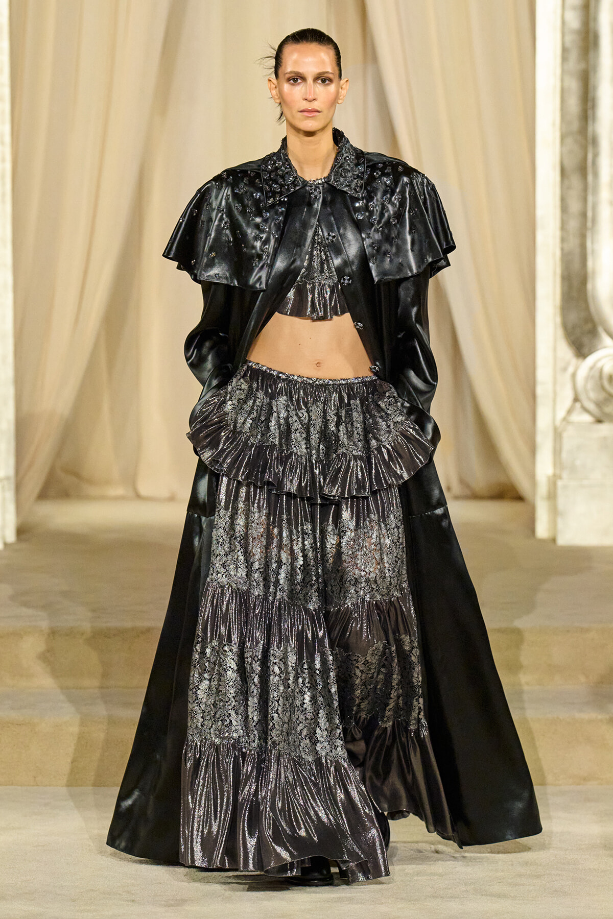 Model walks the runway in a black metallic, tiered gown with lace panels and a dramatic cape-like overlay.