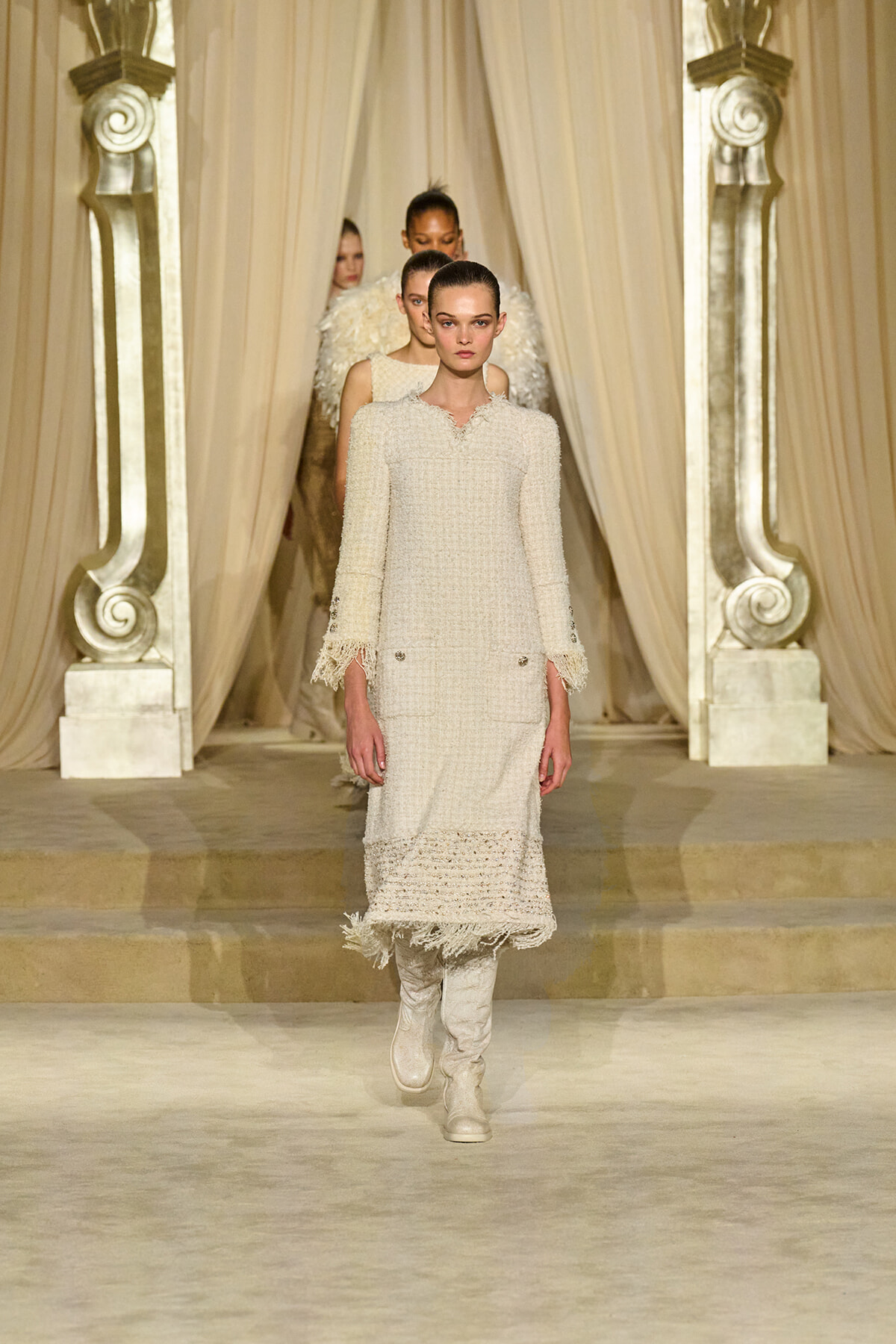Model walking the runway in a cream textured coat dress with fringe cuffs and knee-high boots, ornate golden backdrop behind beige drapes.